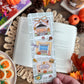 Hand holding a decorative bookmark with autumn-themed illustrations on a book surrounded by fall decorations.