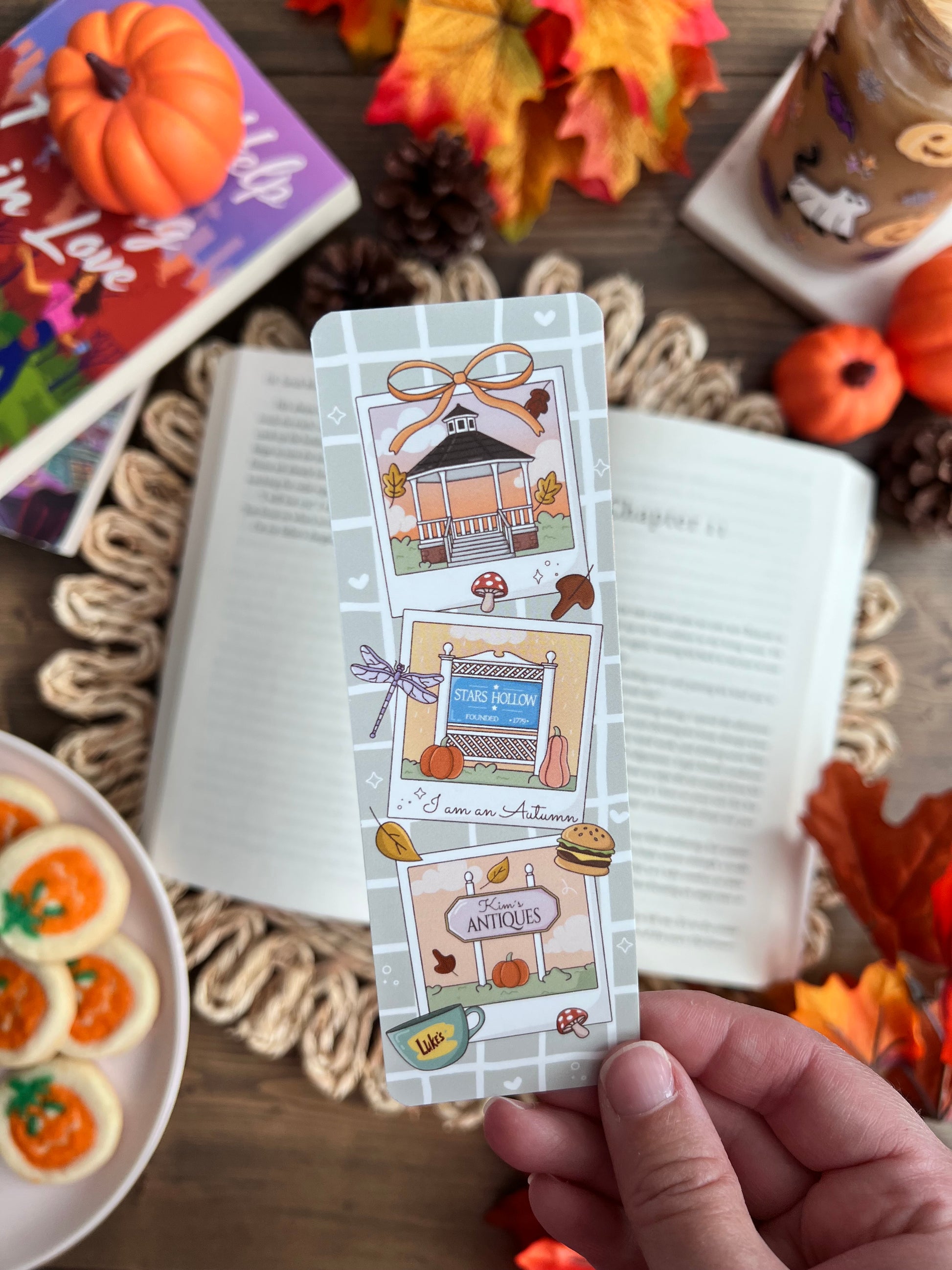 Hand holding a decorative bookmark with autumn-themed illustrations on a book surrounded by fall decorations.